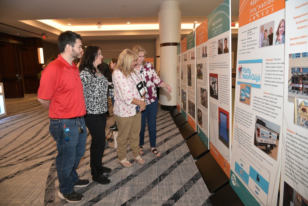 Four people view a series of tall, freestanding event display panels in a conference hallway. The colorful panels showcase case studies and graphics, highlighting professional event signage